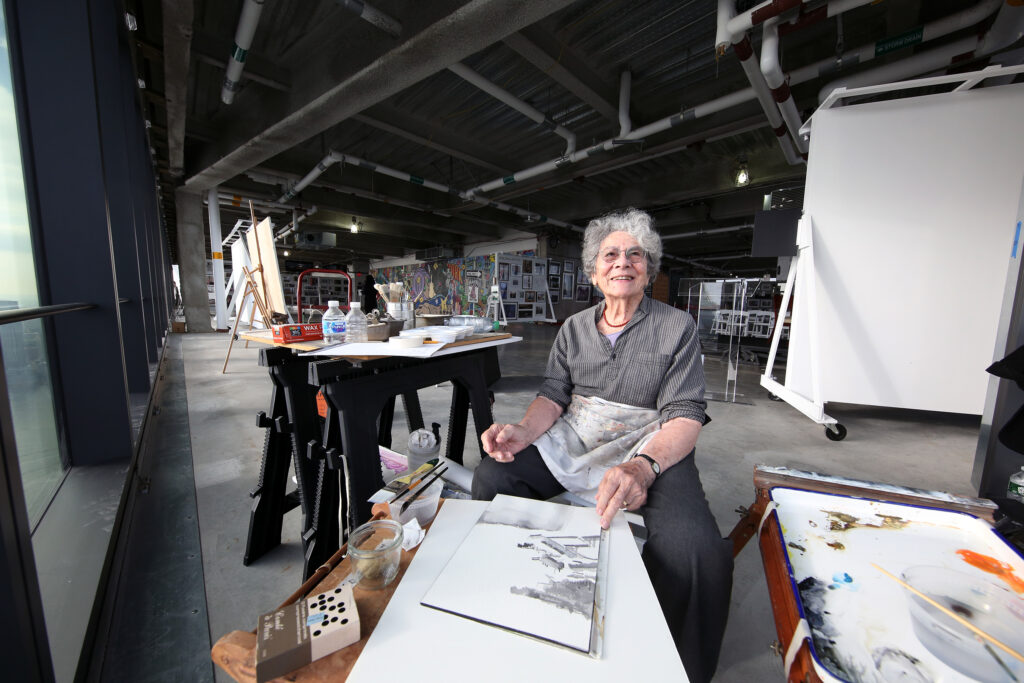 A photo of an elderly white woman sitting in an art studio in an empty office space with high ceilings.